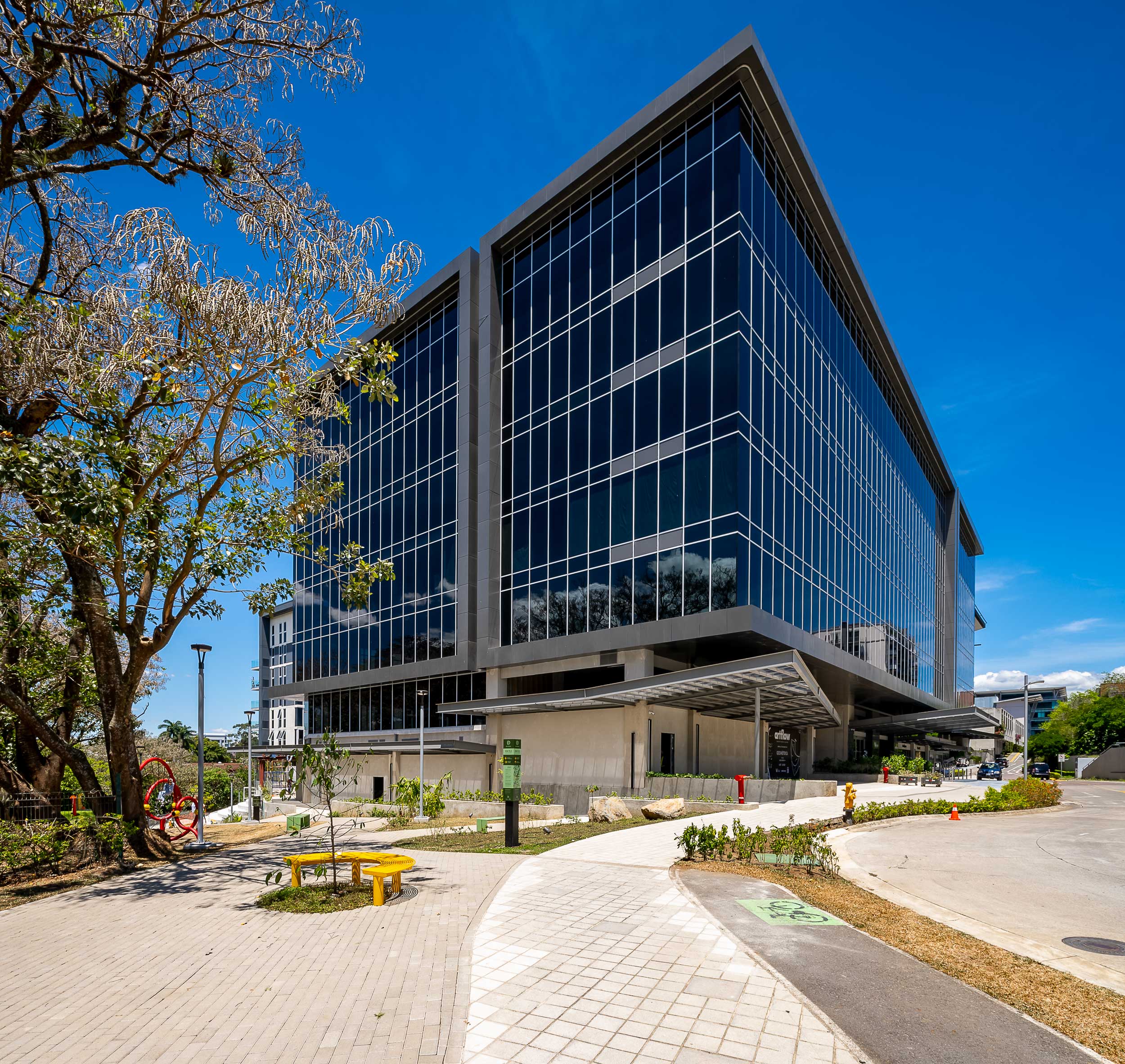 Edificio corporativo moderno de varios pisos con fachada de vidrio y estructura metálica, ubicado en un entorno urbano con aceras amplias, zonas verdes, árboles y mobiliario urbano como bancas y señalización. El cielo está despejado y azul, lo que resalta la arquitectura contemporánea del inmueble