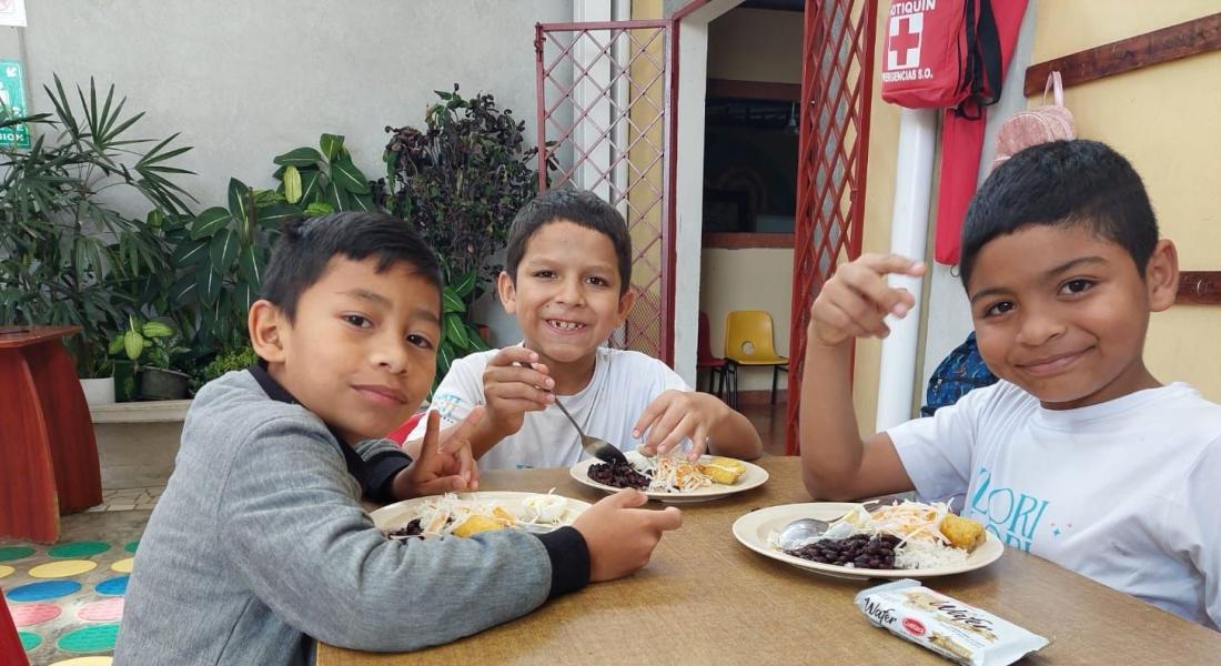 Tres niños están sentados ante una mesa mientras sonríen y comen un plato de comida.
