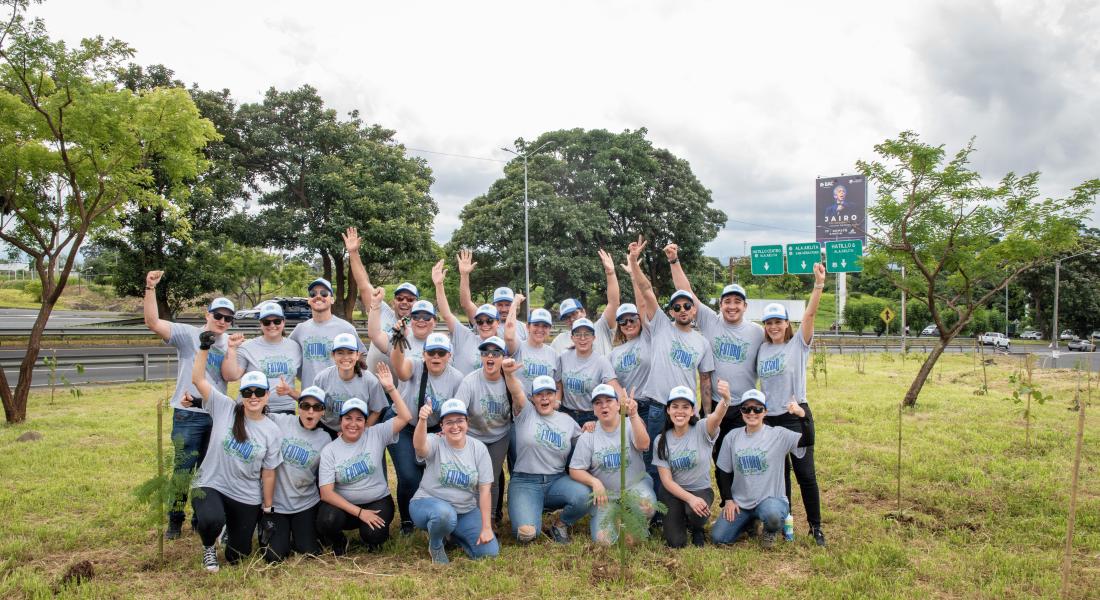 Un grupo de voluntarios vestidos con camisas grises posa en un terreno verde, en medio de pequeños árboles recién plantados.