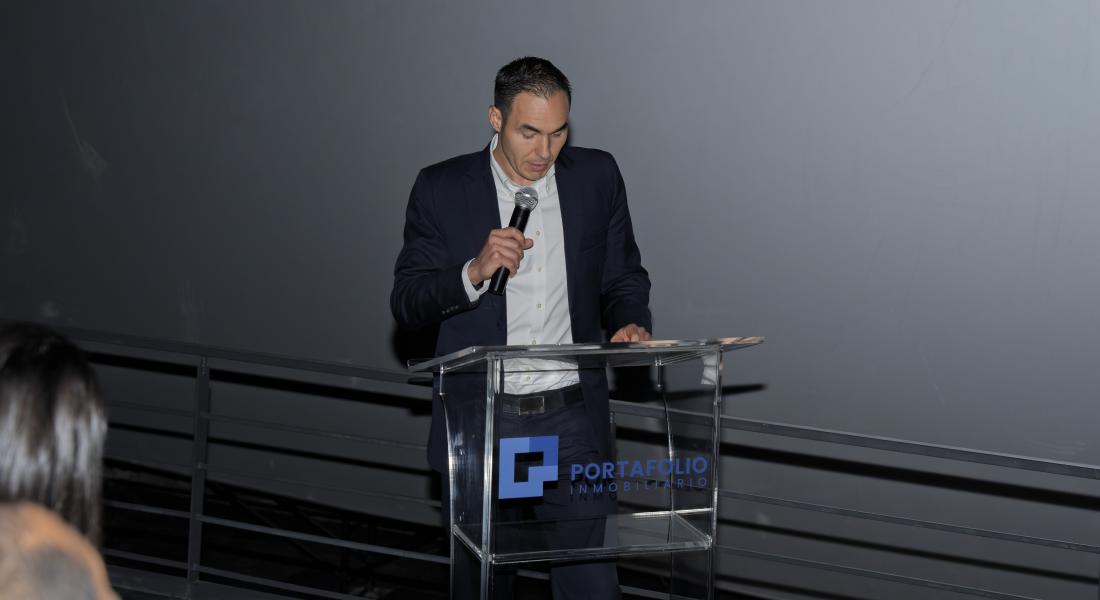 "Alfredo Volio director ejecutivo de la empresa viste de traje oscuro, camisa blanca y corbata se encuentra de pie detrás de un podio transparente con el logotipo de Portafolio Inmobiliario. Está sosteniendo un micrófono mientras parece dirigirse a una audiencia en un evento formal."