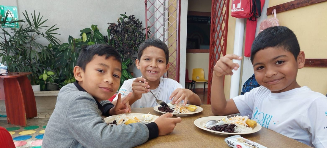 Tres niños están sentados ante una mesa mientras sonríen y comen un plato de comida.
